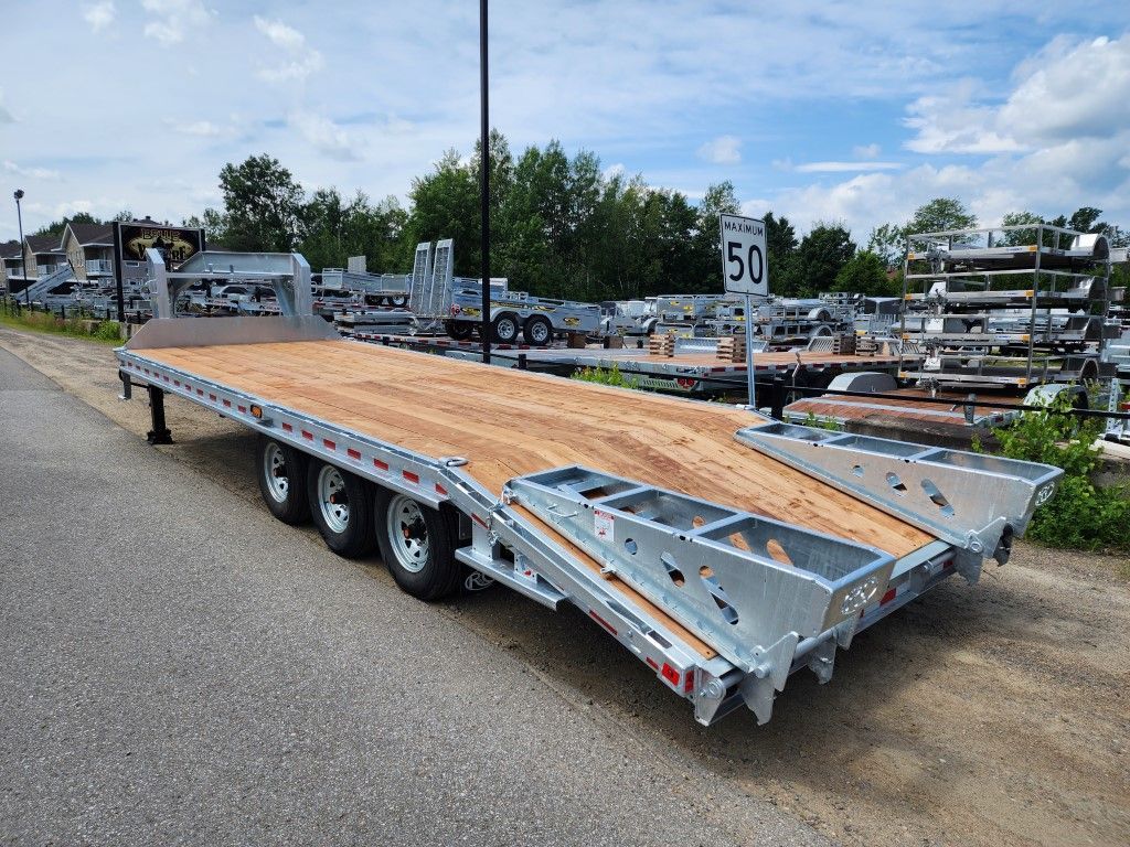 Une grande remorque à plateau avec plancher en bois se trouve sur une route pavée, garée à l'extérieur d'un terrain.