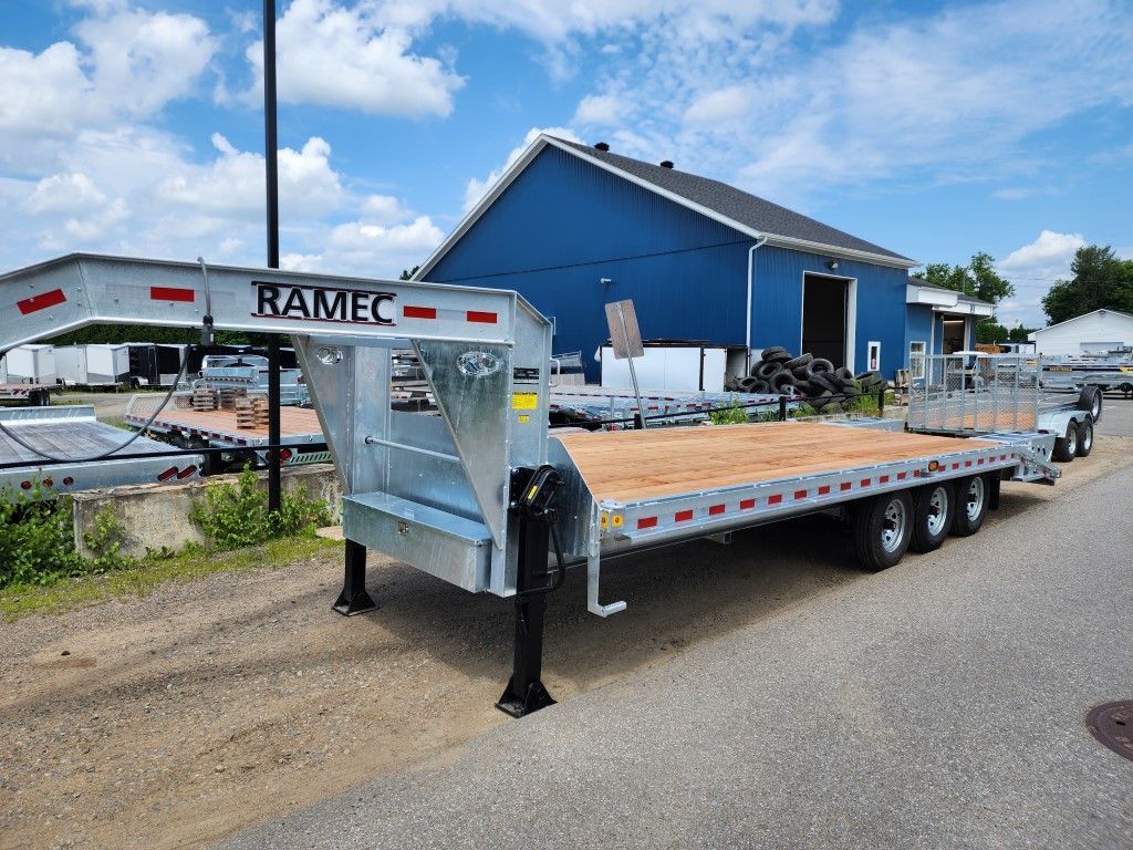 Une remorque à plateau à col de cygne argentée RAMEC garée sur le trottoir près d'un bâtiment bleu par une journée ensoleillée.