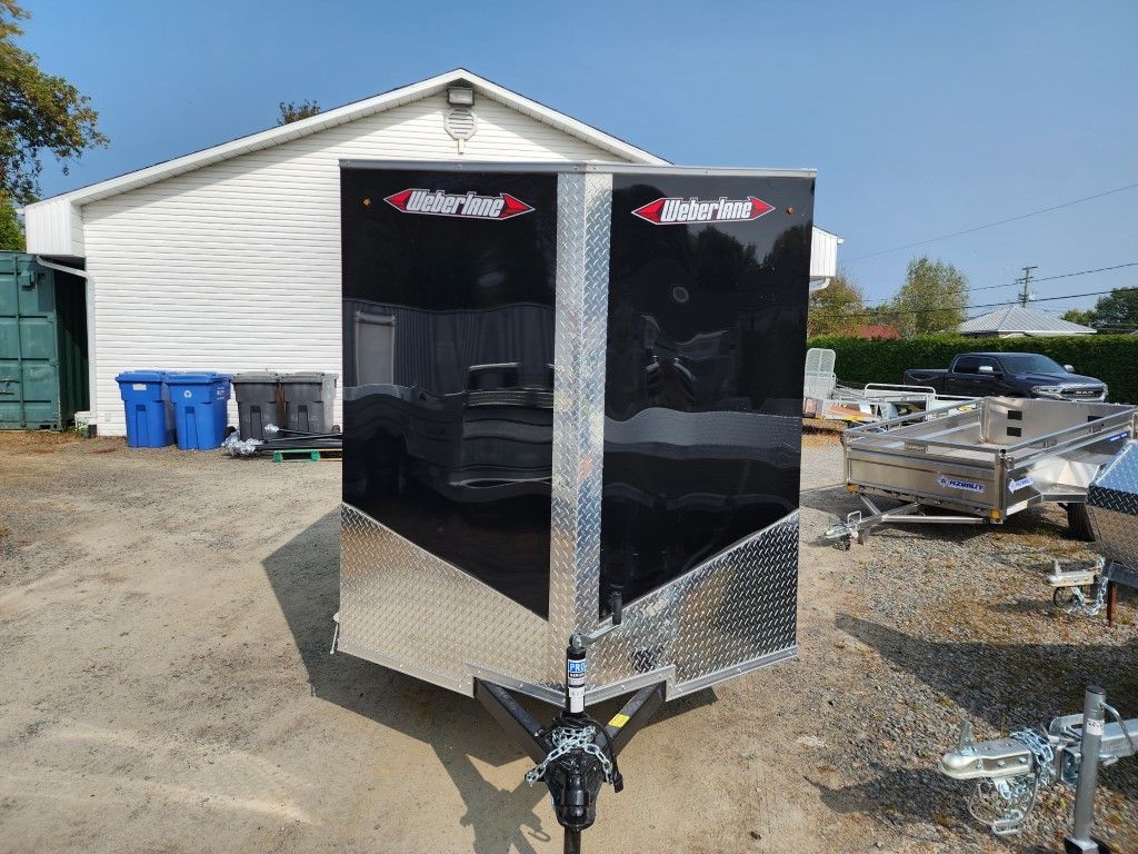 Remorque fermée noire et argentée dans un terrain en gravier, avec un bâtiment blanc en arrière-plan.