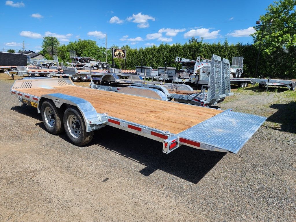 Remorque à plateau avec pont en bois sur un terrain en gravier sous un ciel lumineux.