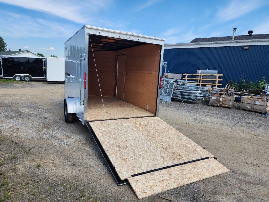 Remorque cargo argentée avec la rampe abaissée, assise sur du gravier devant un bâtiment bleu sous un ciel bleu.