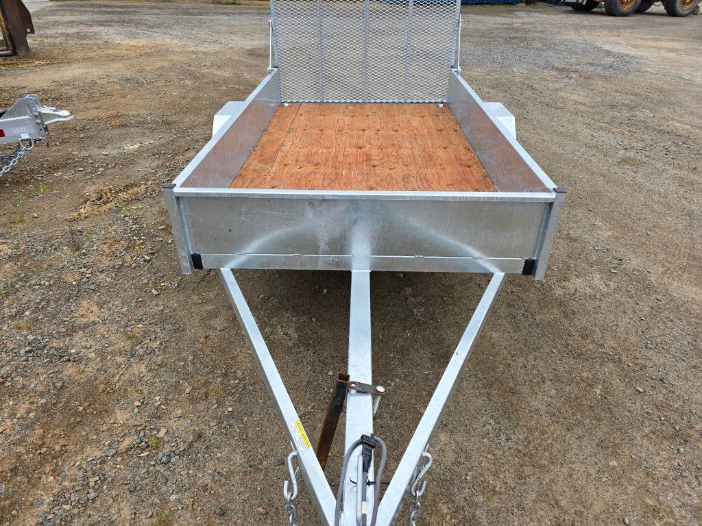 Une remorque utilitaire argentée avec une porte grillagée se trouve à l'extérieur, d'autres remorques et un bâtiment en arrière-plan.