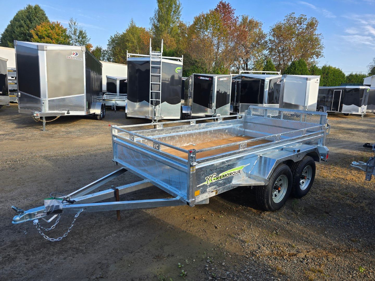 Remorque utilitaire ouverte, couleur argent, avec plancher en bois, garée sur un terrain en gravier. D'autres remorques sont visibles à l'arrière-plan.