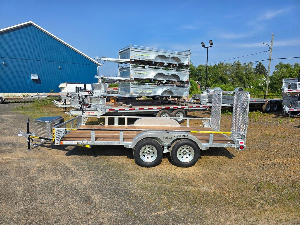 Plusieurs remorques argentées stationnées sur un terrain en gravier ; bâtiment bleu et arbres en arrière-plan.