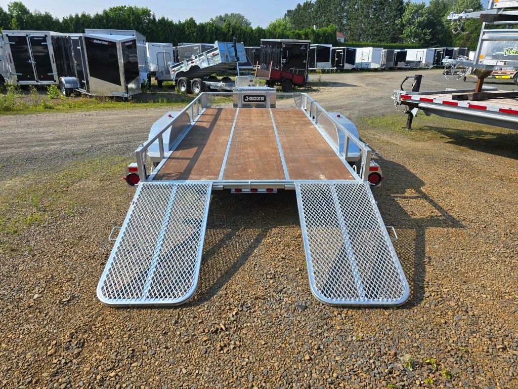 Remorque utilitaire ouverte avec rampes rabattables, garée sur du gravier dans un terrain avec d'autres remorques sous un ciel lumineux.