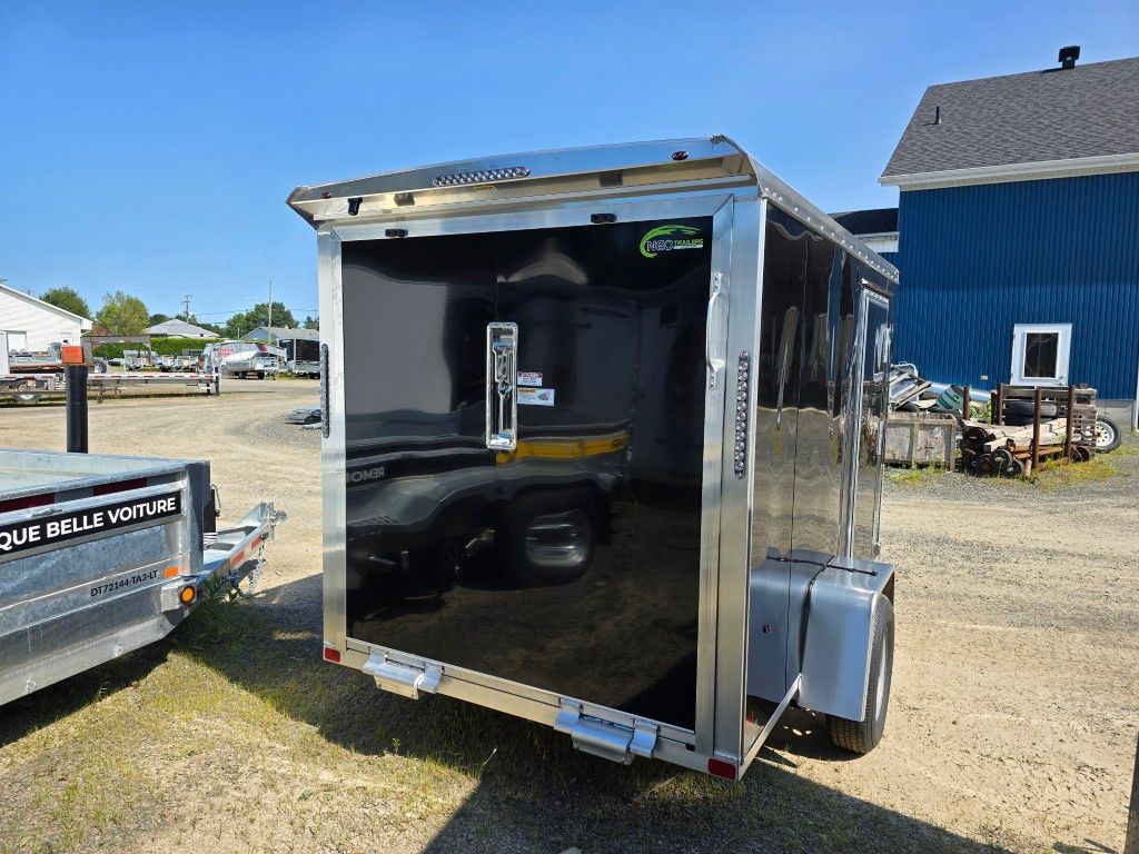 Remorque cargo noire fermée avec garniture argentée, garée sur un terrain en gravier. Journée ensoleillée.