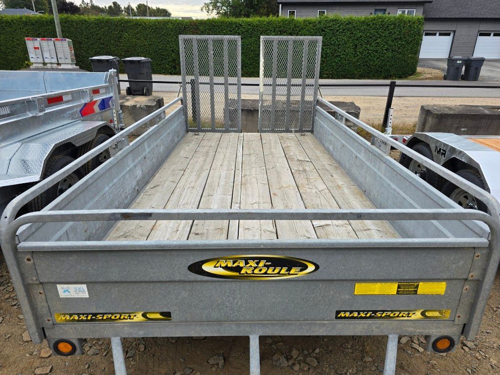 Une remorque utilitaire grise avec un plateau en bois, des rampes et le logo « MAXI-ROULE ».