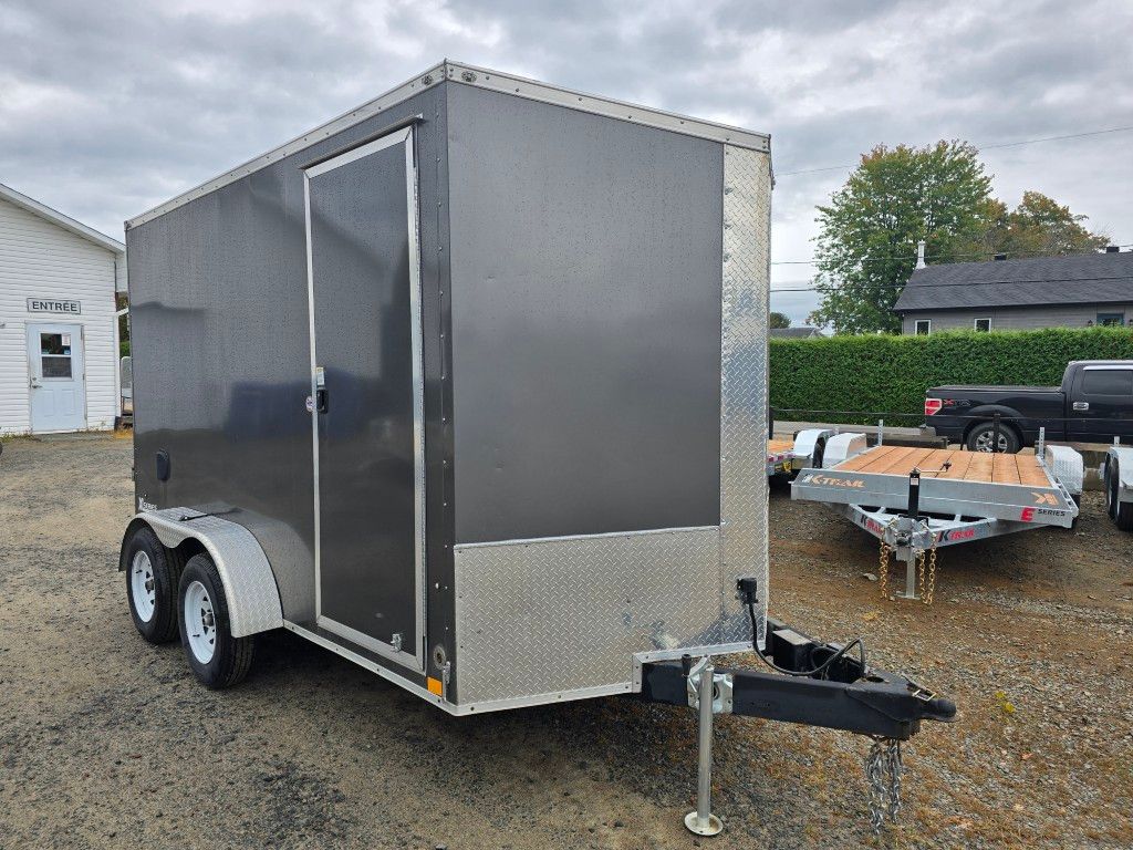 Remorque cargo fermée gris foncé avec roues blanches, garée à l'extérieur sur du gravier.
