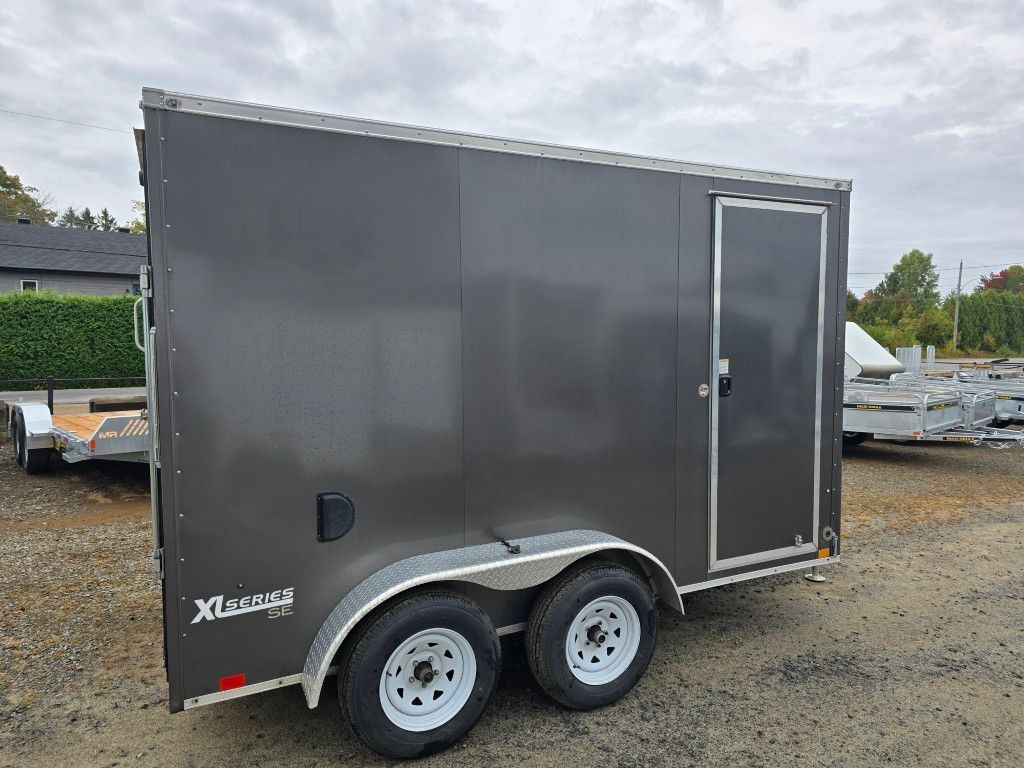 Remorque fermée grise avec roues blanches; stationnée à l'extérieur sur du gravier.