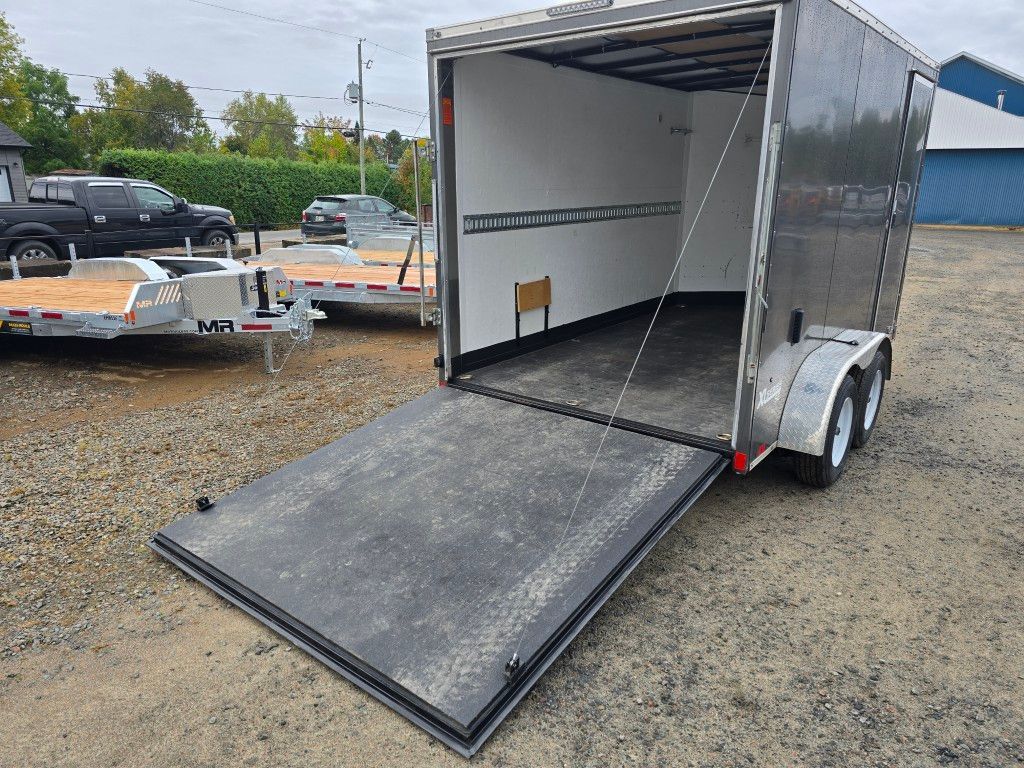 Remorque fermée grise avec rampe ouverte sur terrain en gravier.