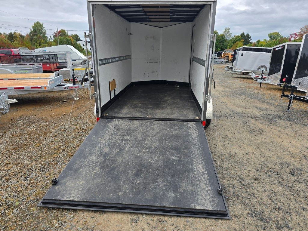 Une remorque cargo blanche vide avec sa rampe ouverte, garée à l'extérieur sur du gravier.