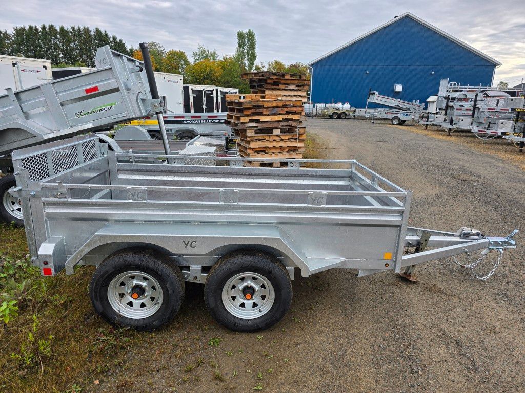 Remorque utilitaire argentée à roues jumelées, stationnée à l'extérieur près d'un bâtiment bleu et de palettes.