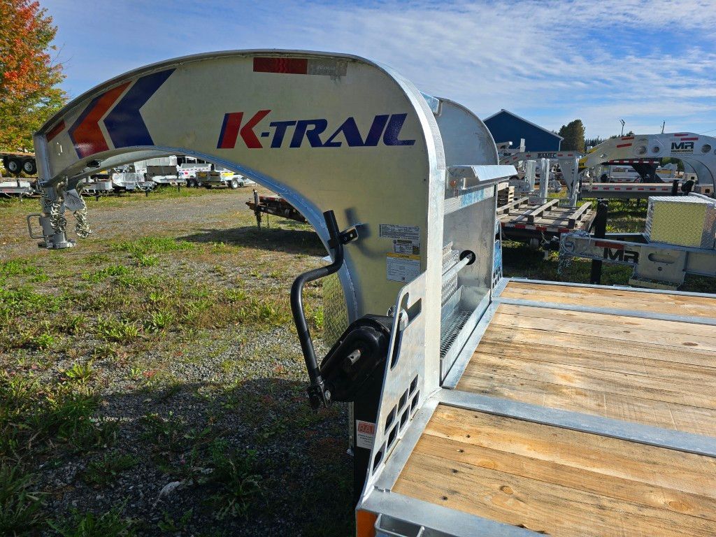 Remorque à plateau stationnée à l'extérieur sur du gravier avec un logo K-Trail blanc et rouge en arrière-plan.
