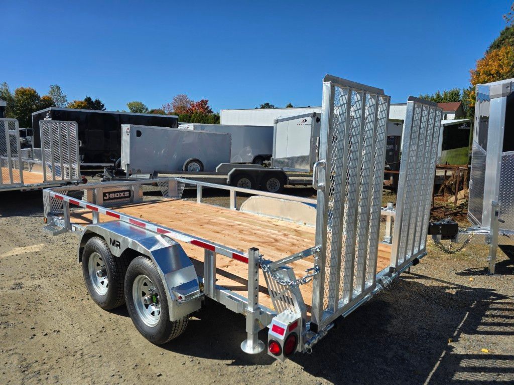 Remorque utilitaire argentée avec pont en bois et rampe en métal, sur terrain en gravier, ciel bleu.