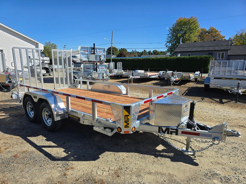 Remorque en aluminium avec pont en bois, coffre à outils et rails latéraux dans un lot avec d'autres remorques sous un ciel bleu.