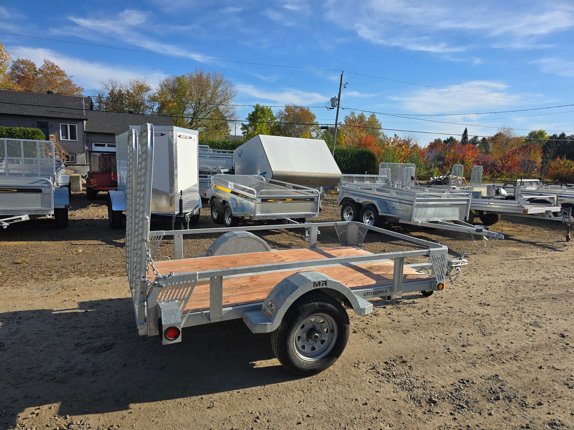 Une petite remorque utilitaire argentée avec un plateau en bois est garée dehors par une journée ensoleillée.