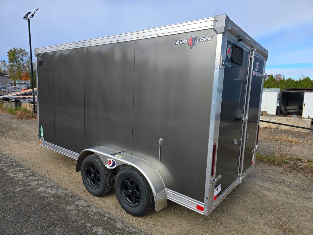 Remorque cargo fermée grise sur gravier. Deux pneus noirs, une porte latérale et des garnitures argentées.