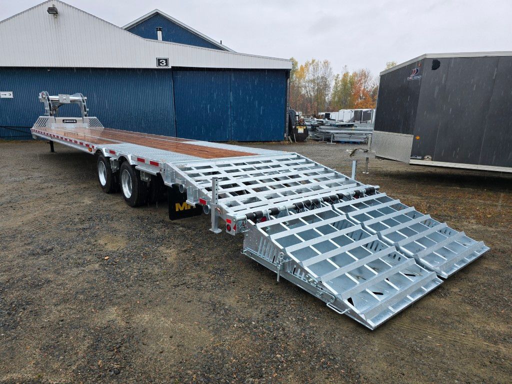 Remorque plateau argentée avec rampes, stationnée devant un bâtiment bleu.