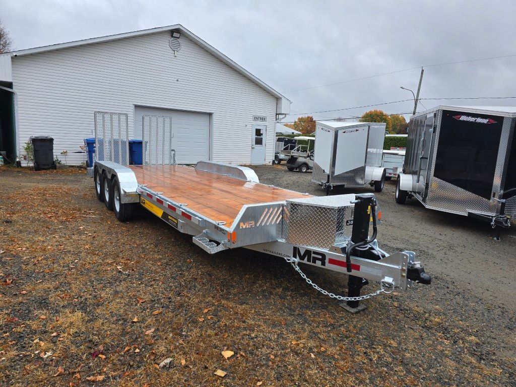 Une remorque à plateau en bois, garée sur du gravier devant un bâtiment blanc.