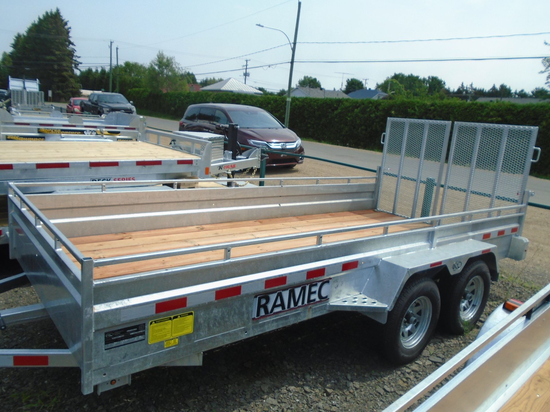 Une remorque utilitaire argentée Ramtec avec pont en bois, deux essieux et garde-corps de chargement, garée à l'extérieur.