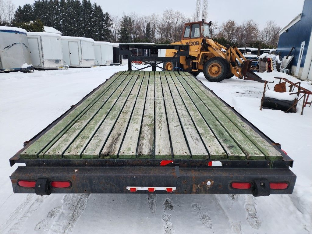 Une remorque à plateau est garée dans la neige avec un tracteur jaune en arrière-plan.
