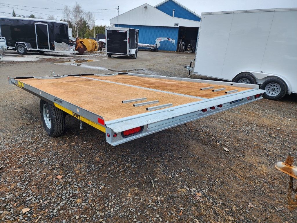 Une remorque à plateau est garée sur un terrain en gravier à côté d'une remorque blanche.