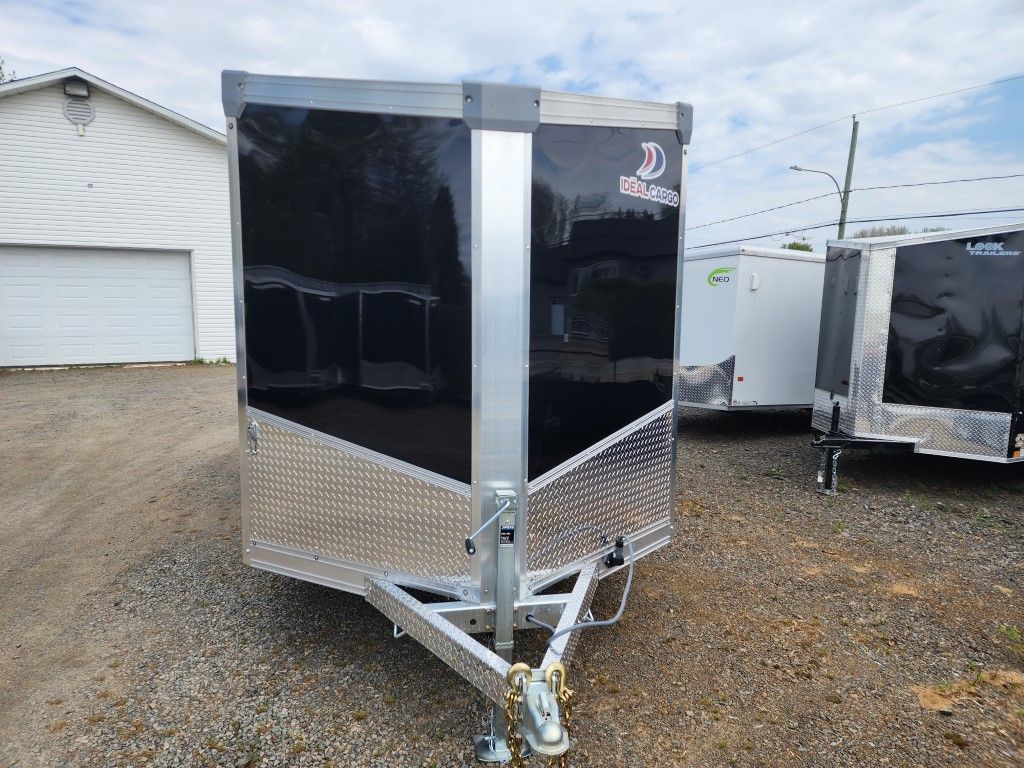 Une remorque noire et argentée est garée sur un terrain en gravier devant un garage.