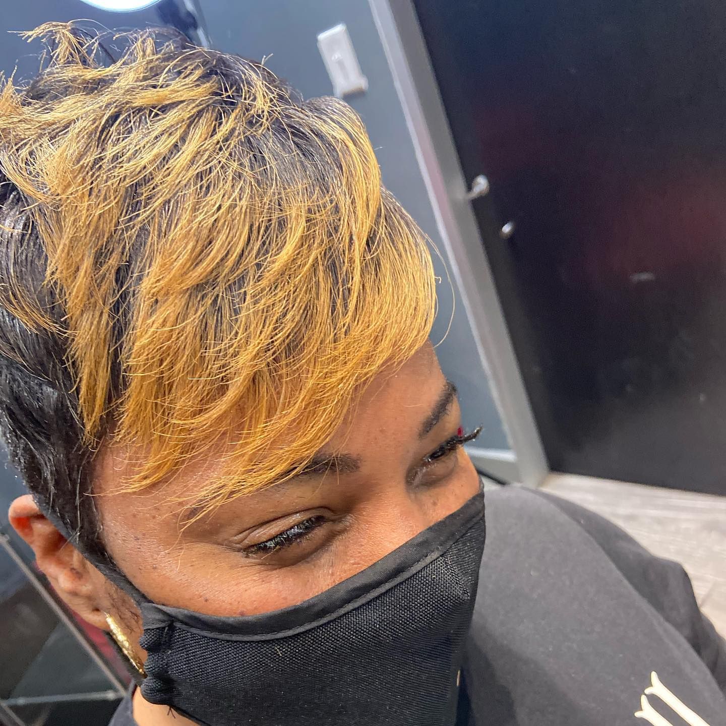 Woman with a short haircut; black roots fade to blonde. Wearing a black mask. Inside a salon.