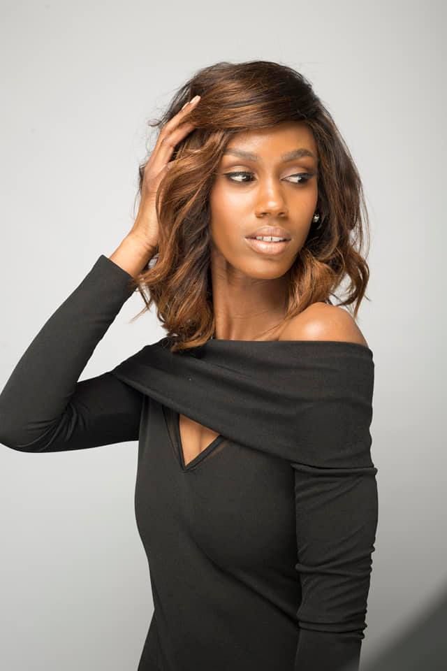 Woman with brown hair in a black off-shoulder dress looks to the side, hand in hair, against a gray backdrop.