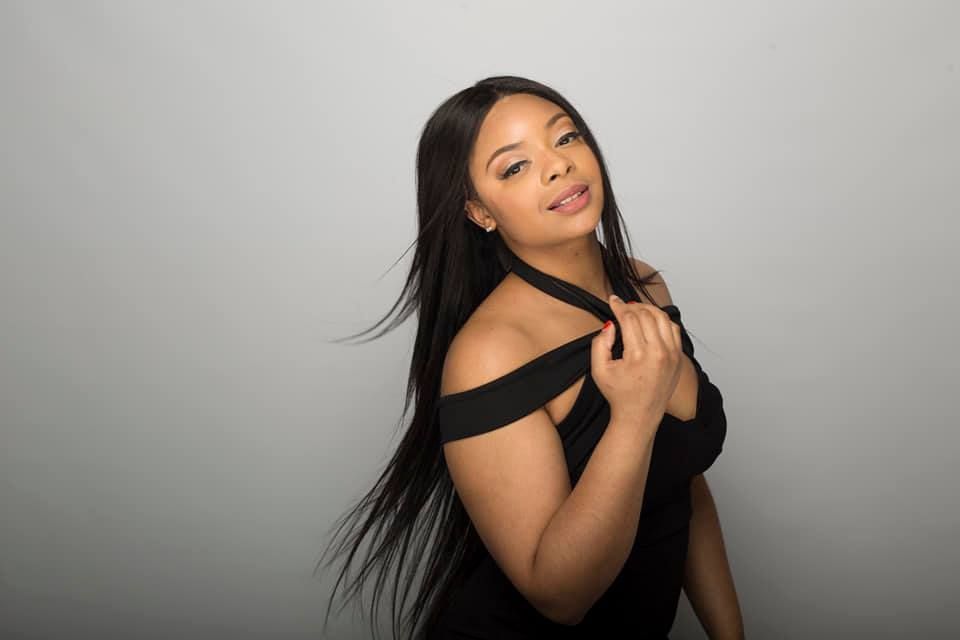 Woman with long black hair in black off-the-shoulder dress, posing against a gray background.