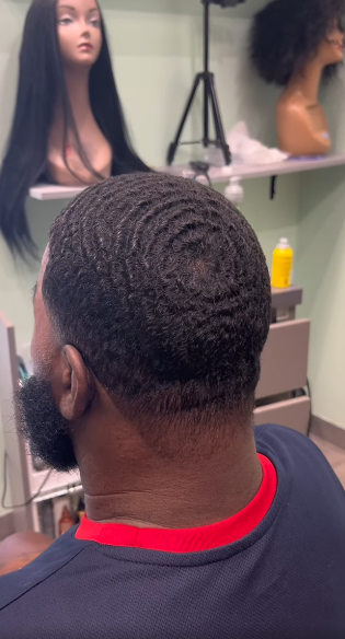 Man with a wave haircut, dark skin, wearing a red and blue shirt, in a hair salon.