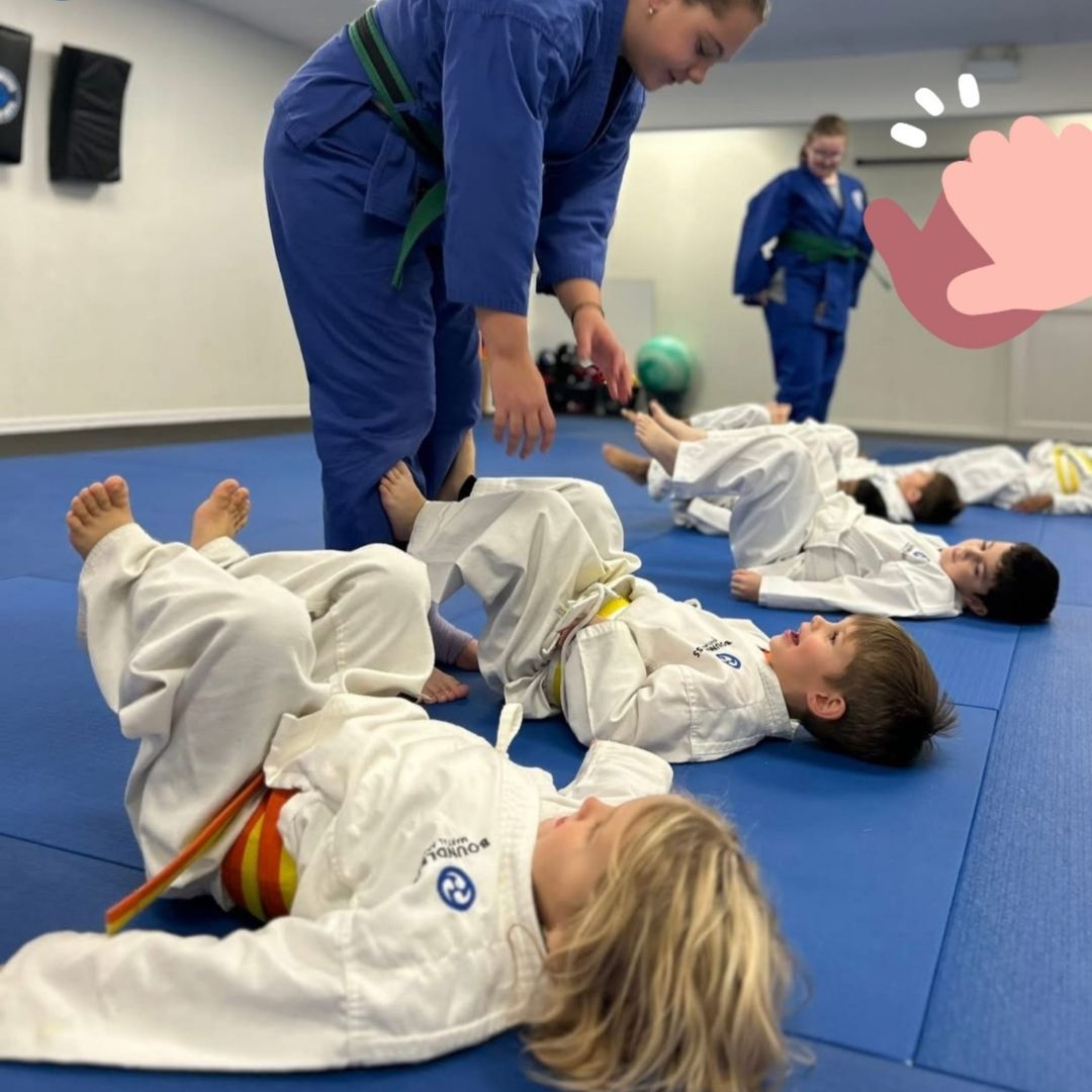 Two children practicing Brazilian Jiu-Jitsu on a mat. One child in white gi has the other in a leg lock. A girl watches.