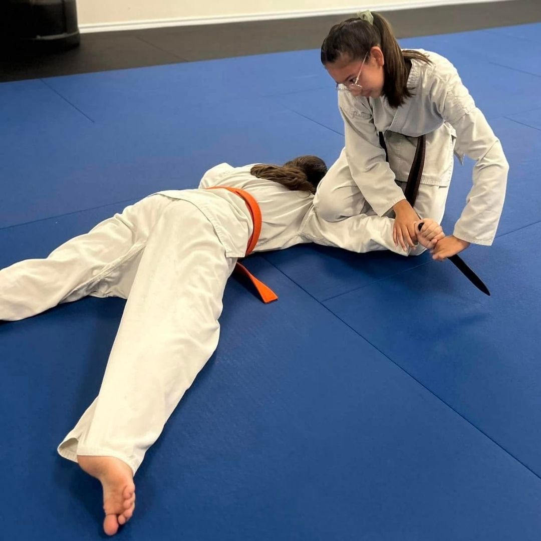 Two men in grappling position on a mat. One embraces the other from behind, both smiling. Gym setting.
