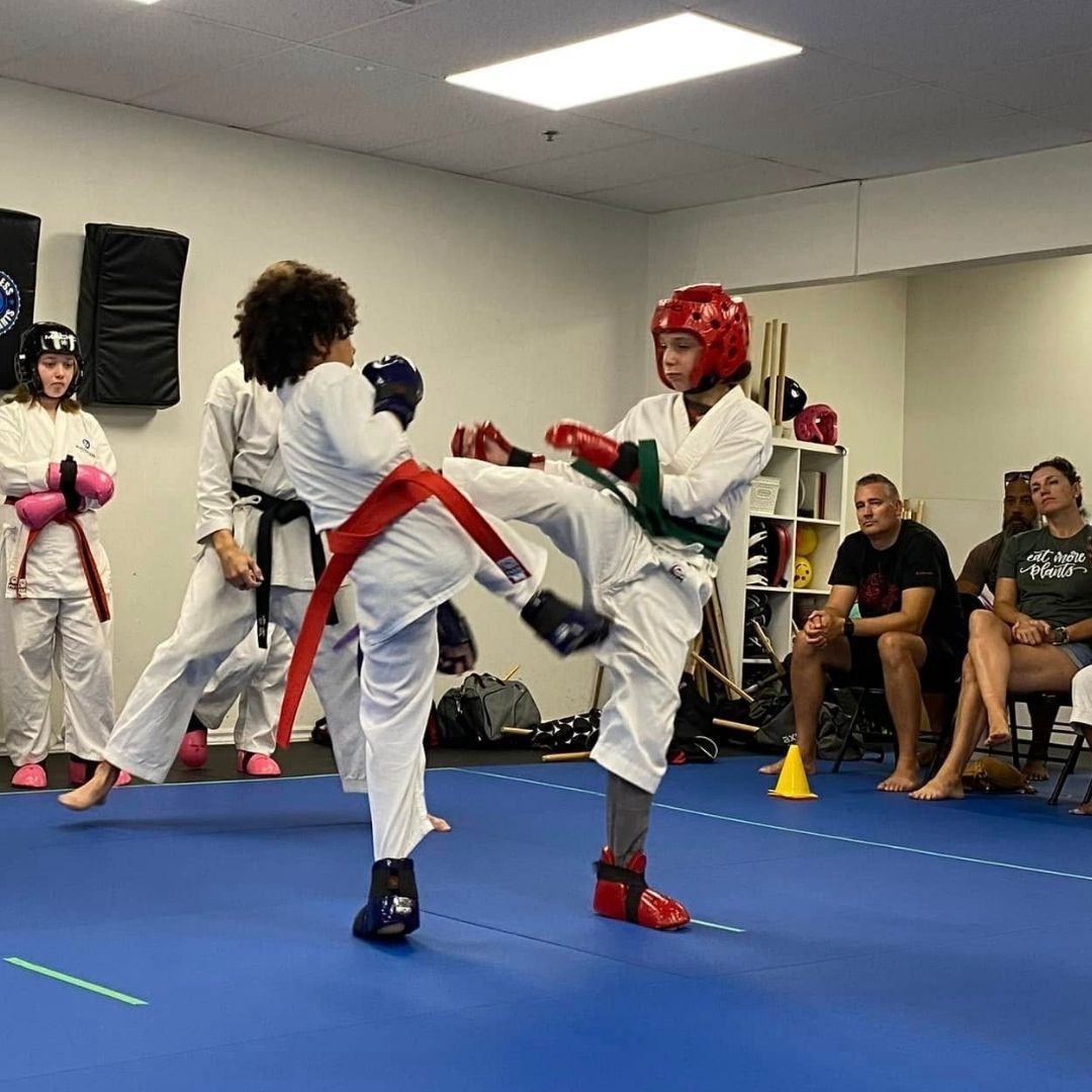 Children in colorful gis play a game on a mat. Two instructors pull a student in blue.