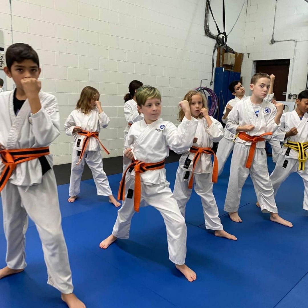 Two young children in blue and black martial arts uniforms grappling on a gym floor, one smiling.