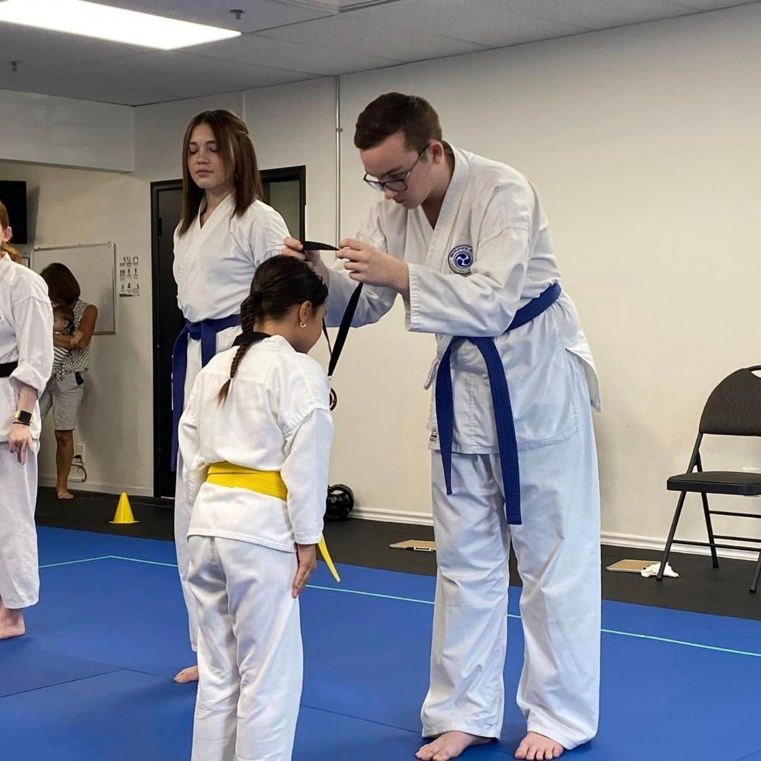 Two children in black and white martial arts uniforms practice a chokehold on a mat. One child smiles.