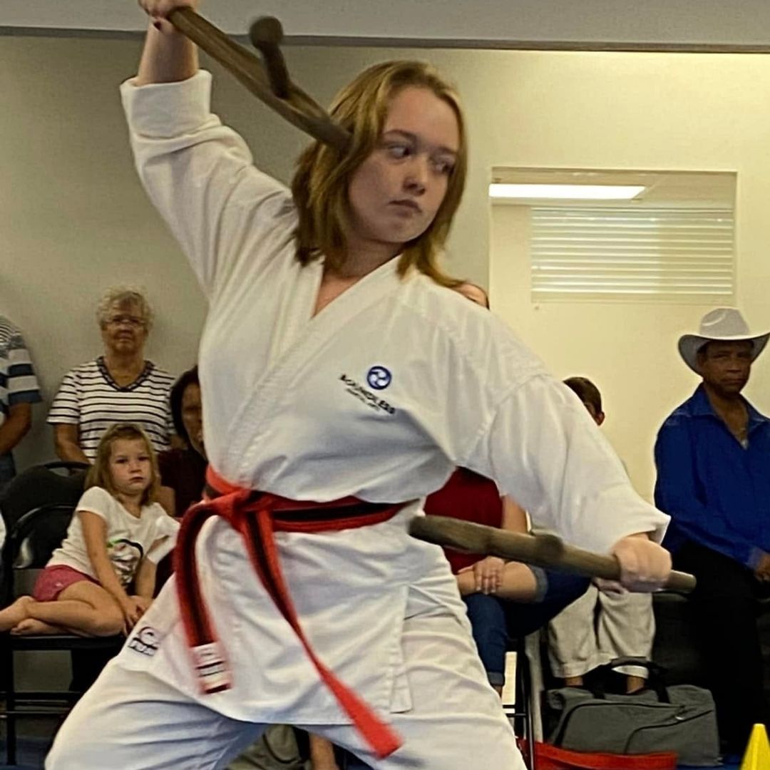 Two men in grappling position on a mat. One embraces the other from behind, both smiling. Gym setting.