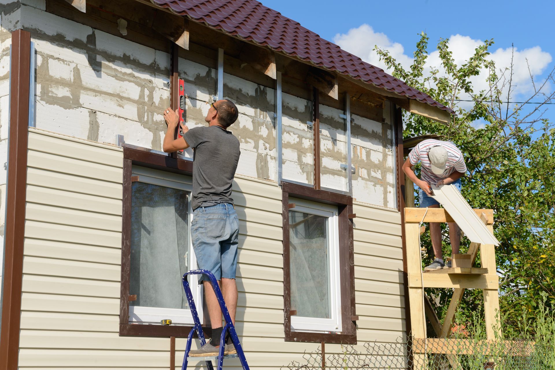 Two people install siding on a house. One uses a level on a ladder, the other holds a panel.