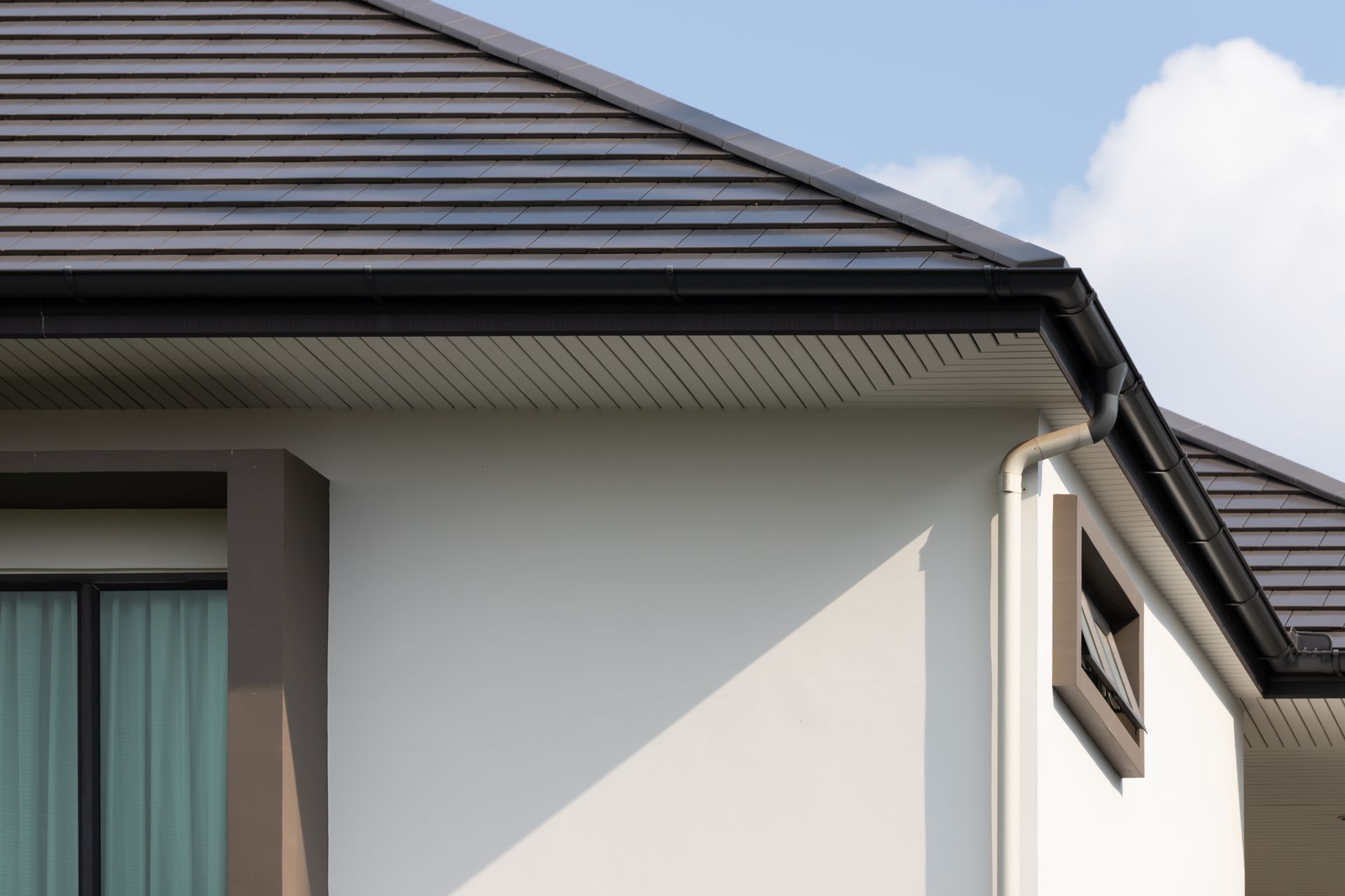 Corner of a modern house with dark roof and black gutters against a blue sky.