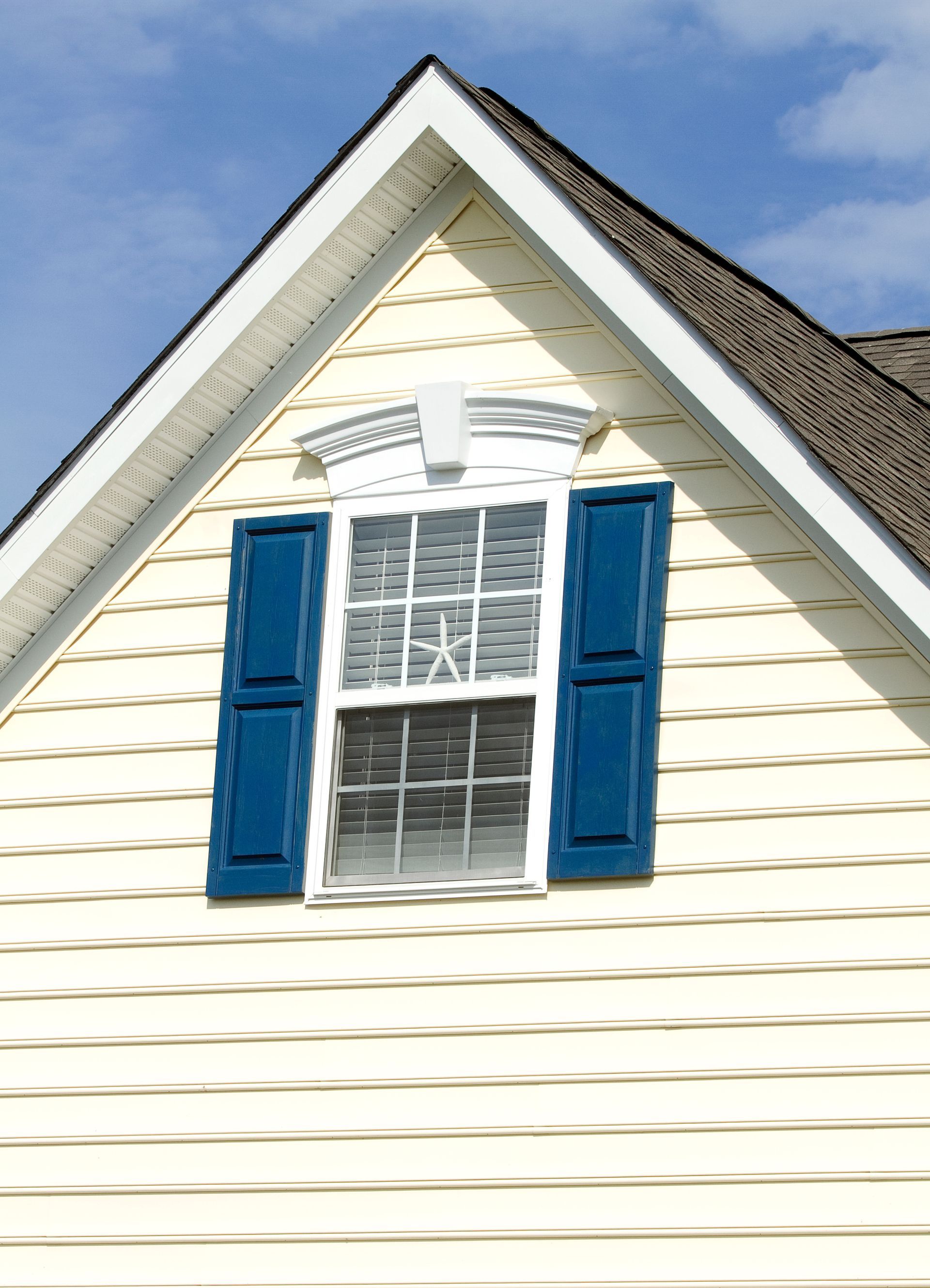 Gabled house exterior with a window, blue shutters, and yellow siding against a blue sky.