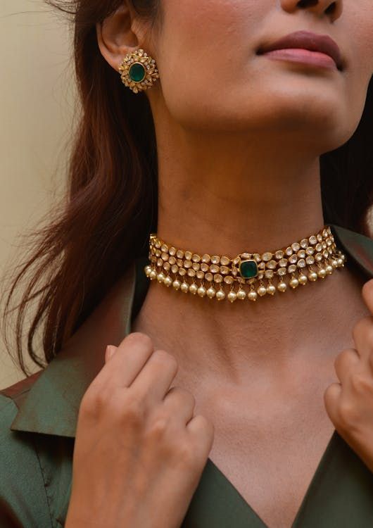 Woman wearing a gold choker necklace with emerald and pearl accents, matching earrings, and green top.