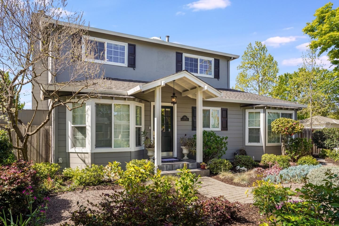 A large gray house with a porch and lots of windows surrounded by trees and bushes.