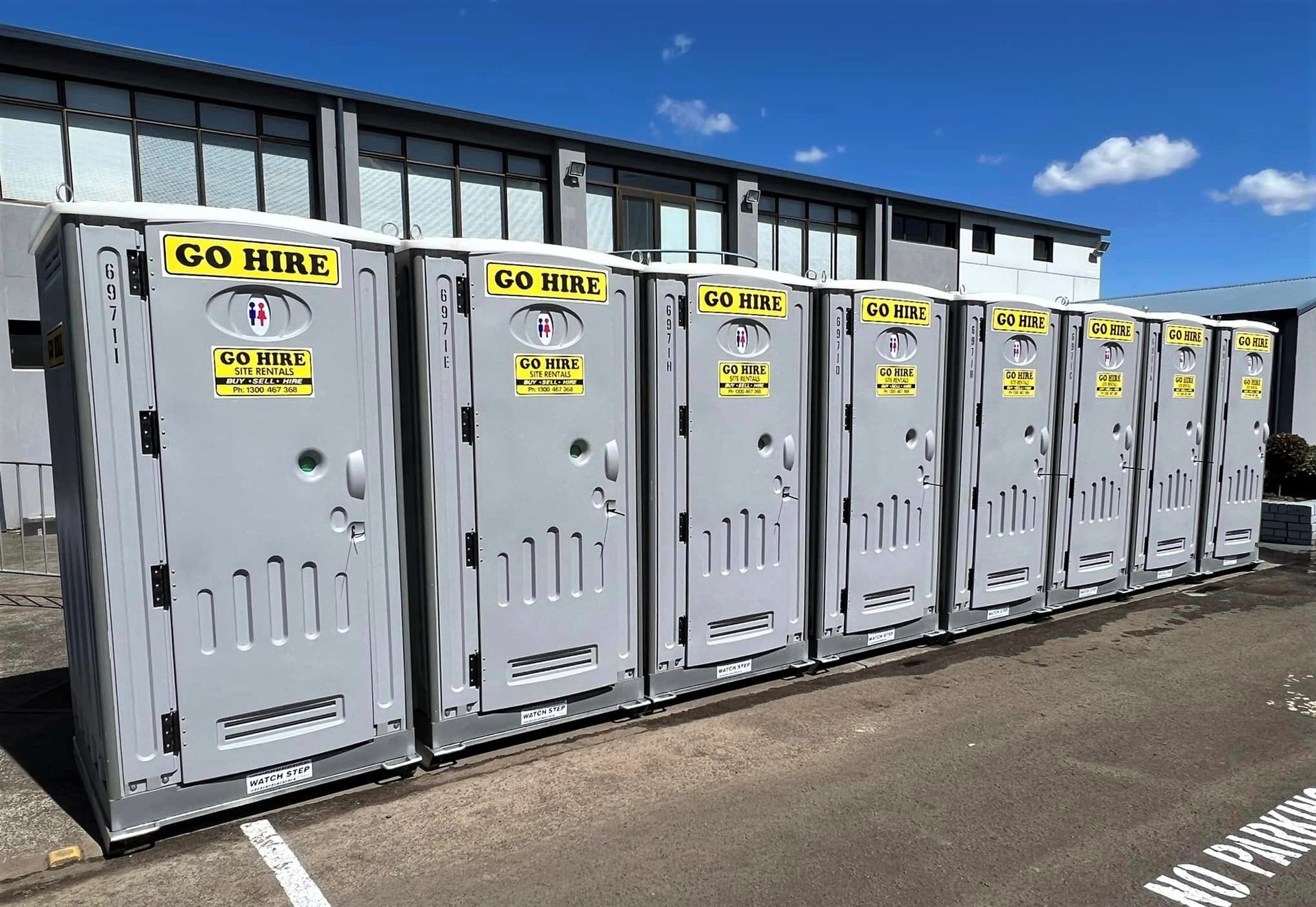 Row of grey portable restroom units lined up outside a commercial building under a clear blue sky.