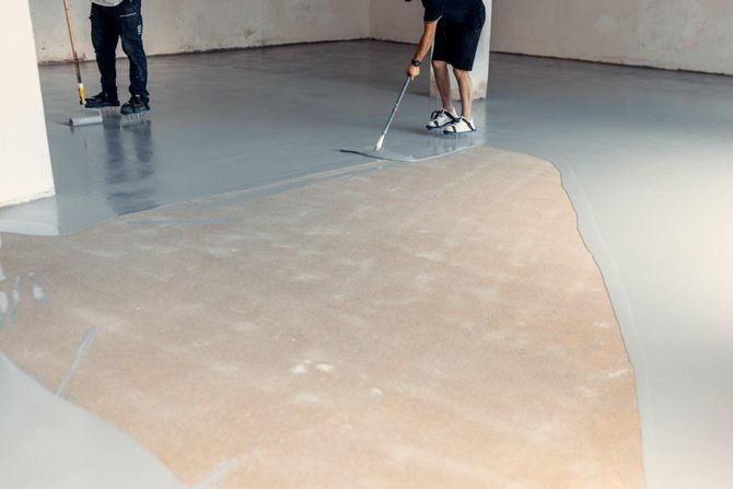 Workers spreading a beige coating across a concrete floor in an unfinished indoor space