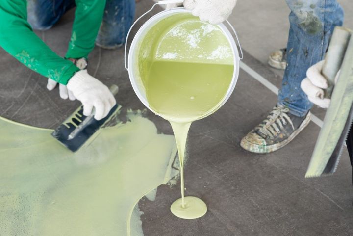 Workers pouring pale green liquid from a bucket onto a concrete floor