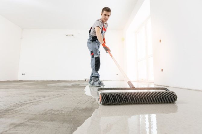 Worker leveling wet concrete with a roller in a bright, empty room