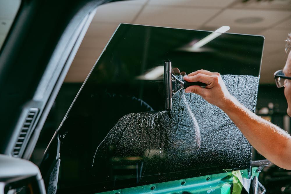 Person applying window tint to a car window with a squeegee inside a workshop.