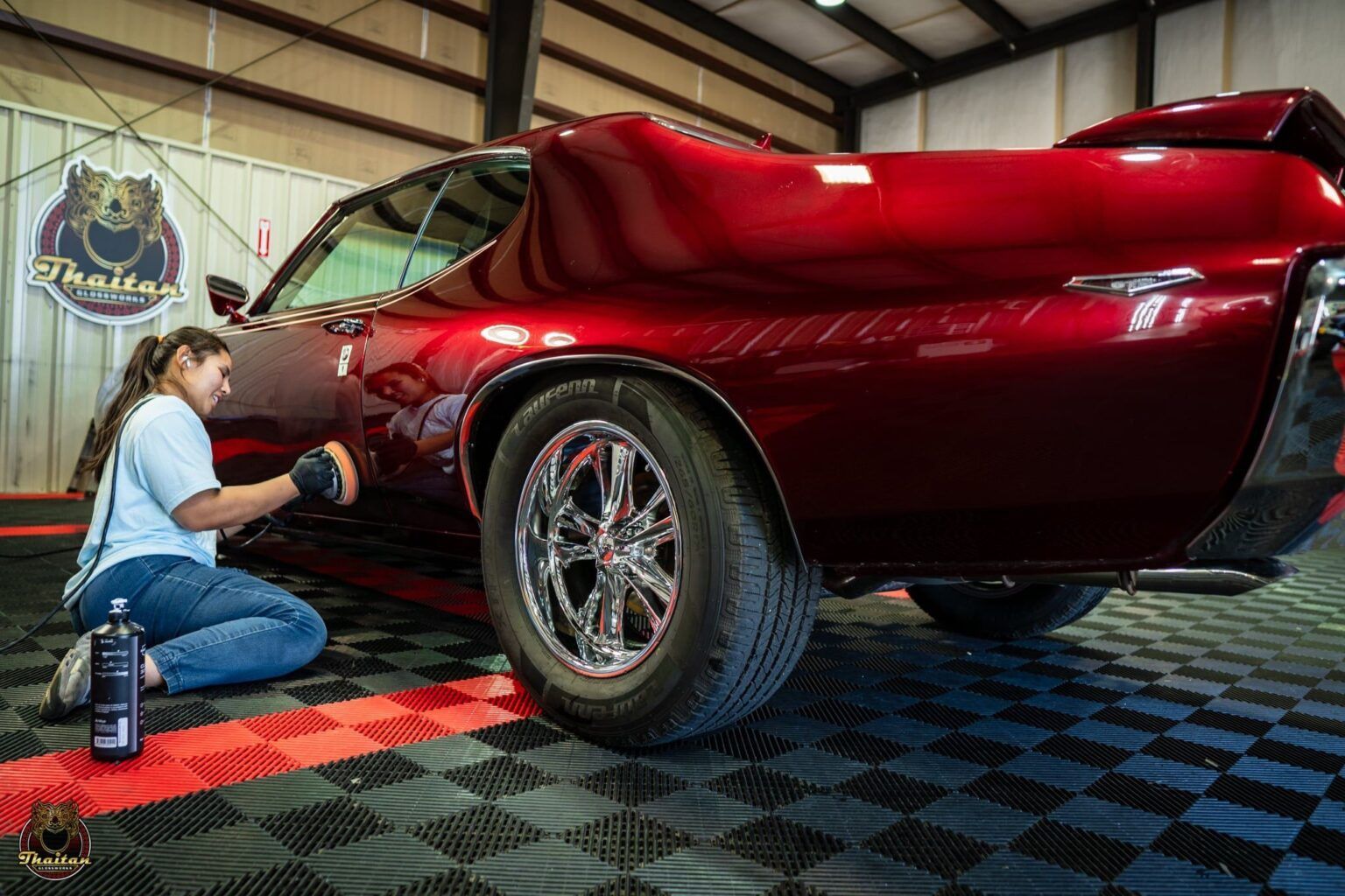 A woman is kneeling down to polish a red car in a garage.