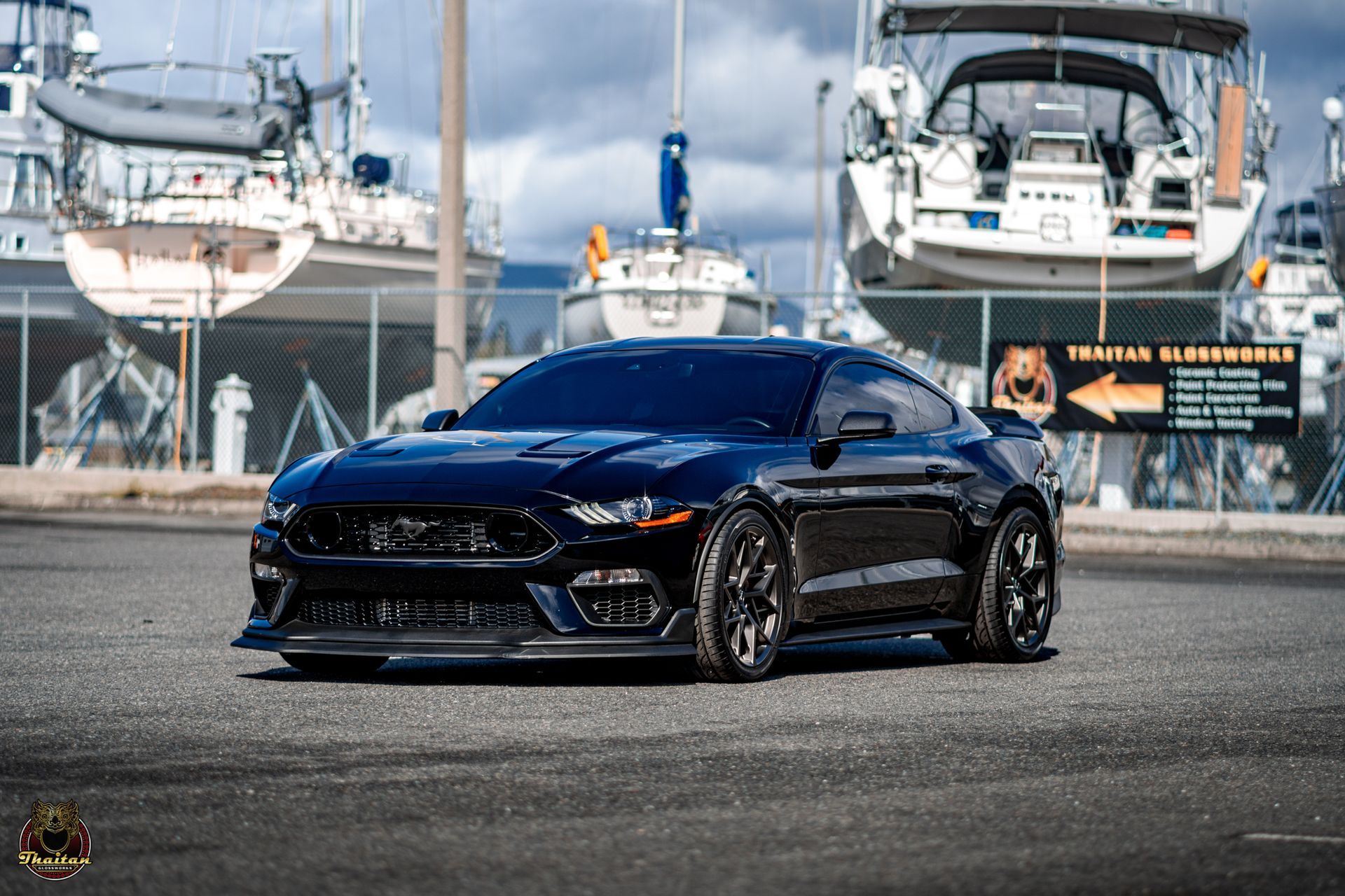 A black mustang is parked in front of a marina with boats.