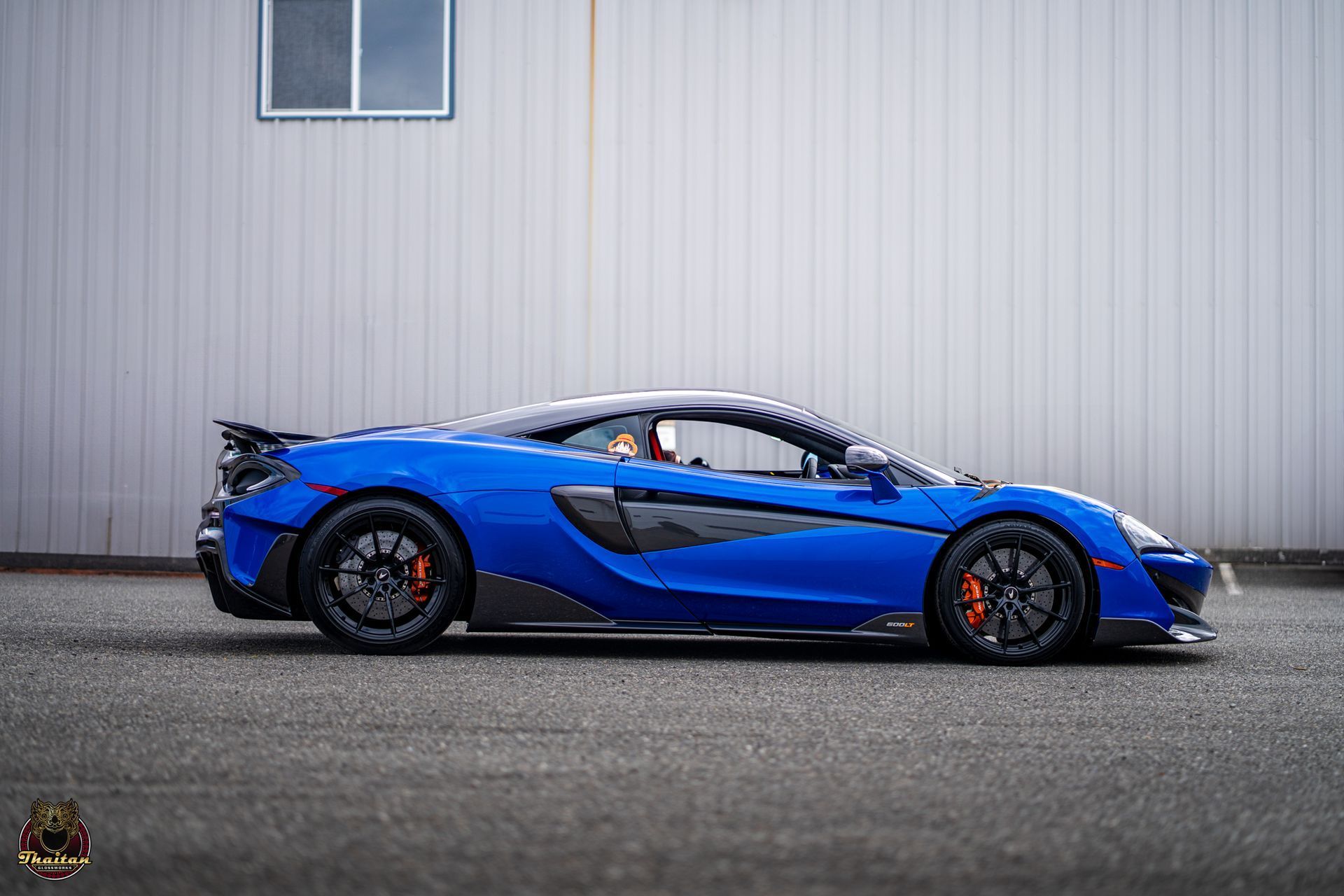 Blue McLaren sports car parked in front of a grey building, black wheels with orange brake calipers.